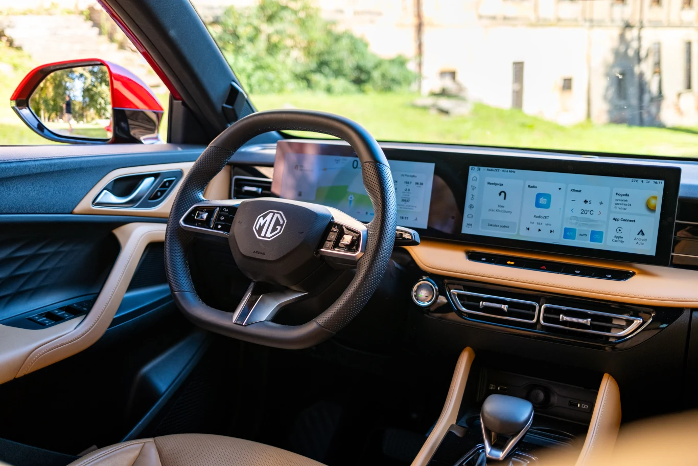 Wnętrze nowoczesnego samochodu marki MG z widoczną kierownicą, ekranem dotykowym na desce rozdzielczej oraz eleganckimi elementami wykończenia w beżowych i czarnych kolorach.