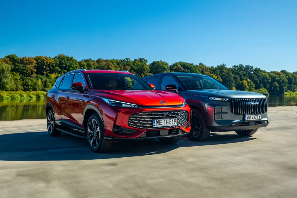 Dwa nowoczesne samochody SUV zaparkowane obok siebie na otwartej przestrzeni przy jeziorze, w tle drzewa i niebieskie niebo.