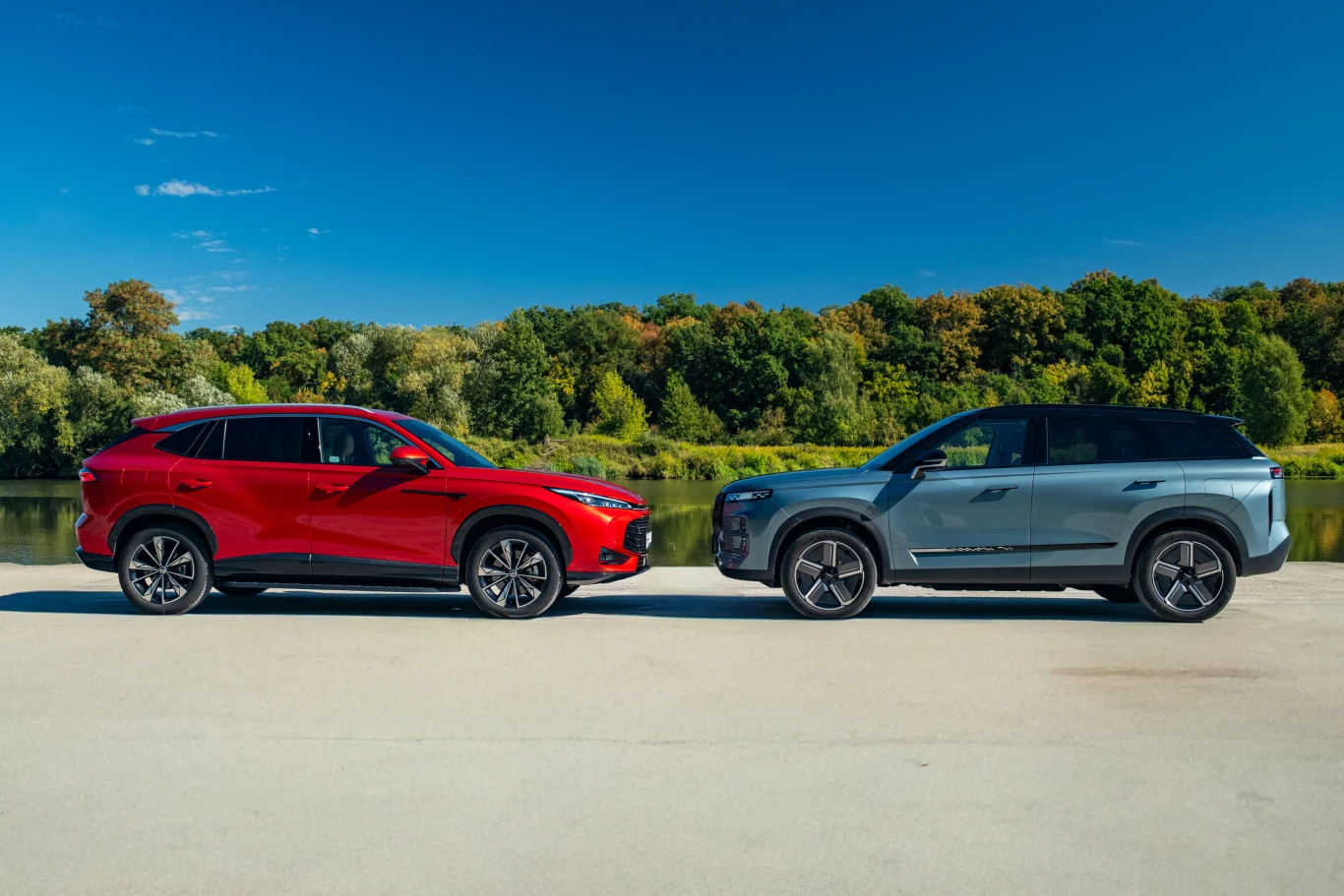 Dwa nowoczesne SUV-y zaparkowane naprzeciwko siebie na tle zielonych drzew oraz spokojnej rzeki, czerwony pojazd po lewej i niebieskoszary po prawej stronie, jasna nawierzchnia oraz bezchmurne, niebieskie niebo.
