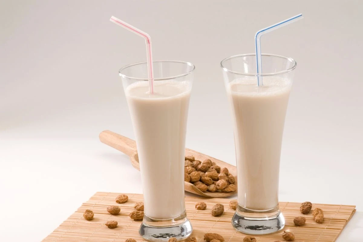 Dwa wysokie szklanki z białym napojem, każda ze słomką w innym kolorze, ustawione na drewnianej podkładce, obok rozsypane orzeszki ziemne lub tygrysie orzechy z drewnianą łopatką w tle.