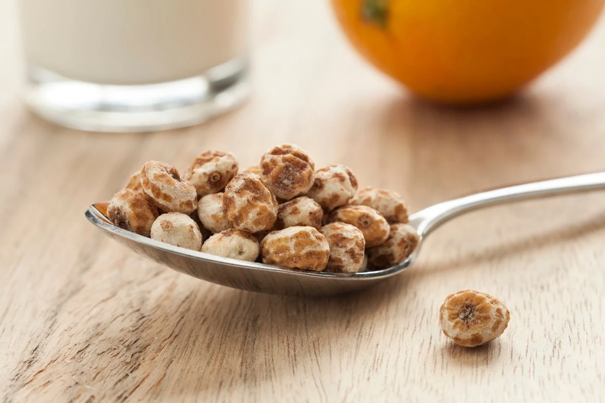 Łyżka pełna chrupiących kulek płatków śniadaniowych umieszczona na drewnianym blacie, w tle szklanka mleka oraz fragment pomarańczy.