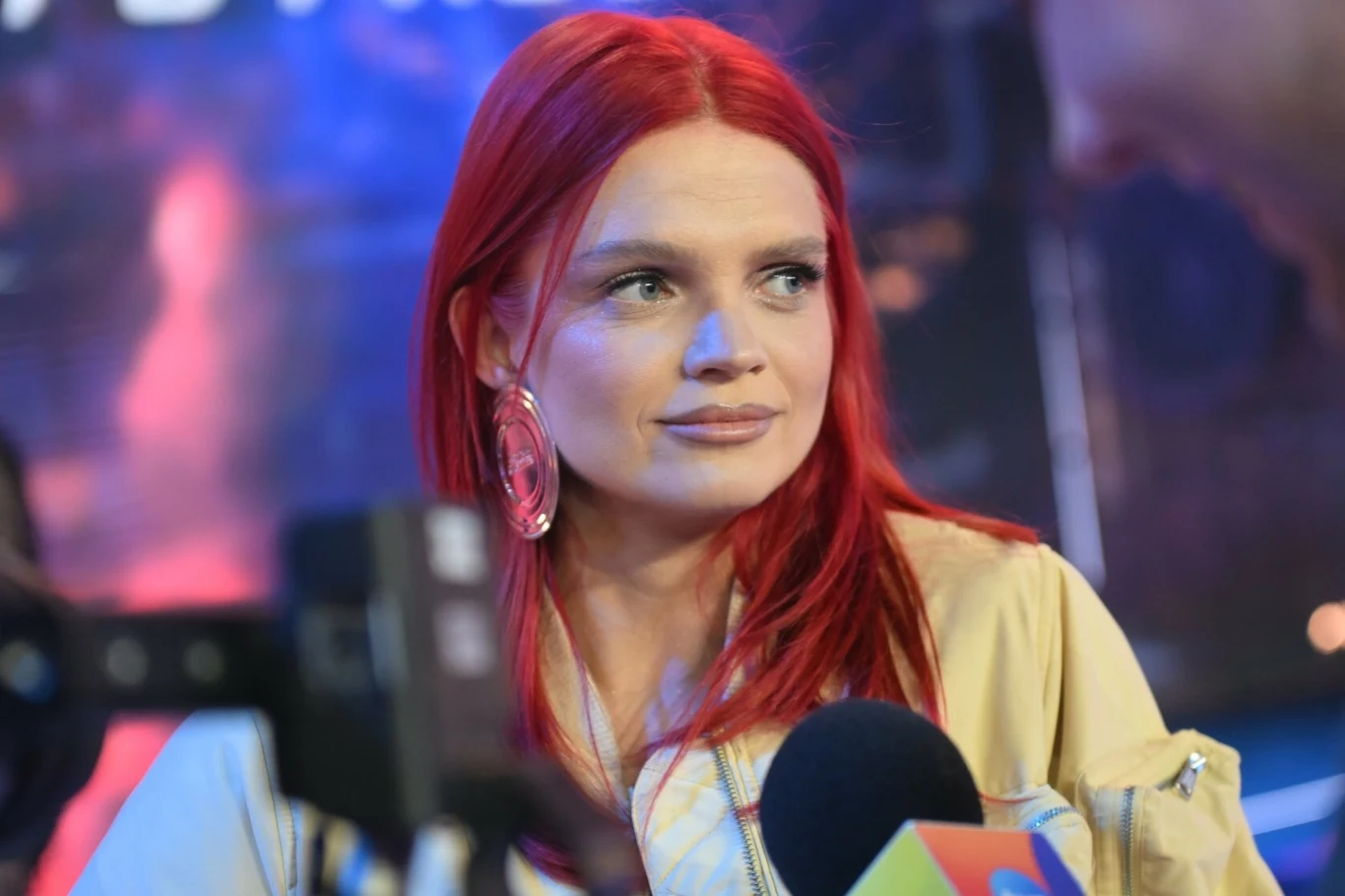 Margaret opowie o swoich wpadkach scenicznych kobieta z intensywnie czerwonymi włosami udziela wywiadu przed kamerą i mikrofonami, w tle rozmyte światła i sylwetki sugerujące medialne lub eventowe otoczenie