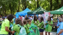 Grupa młodych osób ubranych w zielone koszulki przygotowuje się do akcji sprzątania parku, trzymając worki i rękawiczki, w tle widać namioty organizatorów ustawione wśród zielonych drzew.