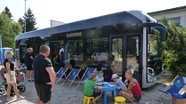 Grupa dzieci i dorosłych siedzi przy stoliku z kolorowankami obok nowoczesnego autobusu miejskiego, w tle rozstawiono leżaki, a całość odbywa się na świeżym powietrzu w słoneczny dzień.