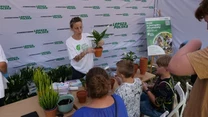 Instruktorka prowadzi warsztaty sadzenia roślin dla dzieci, na stole leżą doniczki, rośliny i ziemia. W tle widoczne stoisko i banery promujące ekologiczną inicjatywę.