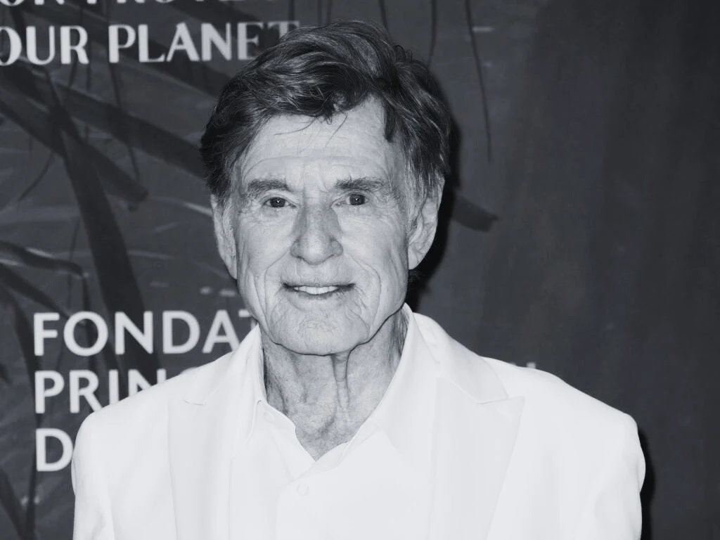 Robert Redford nie żyje. Artysta miał 89 lat Starszy mężczyzna w białej marynarce, uśmiechnięty, stoi na tle ściany z napisem i dekoracjami.