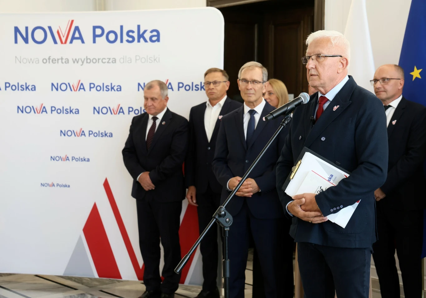 Konferencja założycielska partii Nowa Polska Grupa elegancko ubranych mężczyzn podczas konferencji prasowej przy mikrofonie z planszą Nowa Polska w tle, na której widoczne są biało-czerwone elementy oraz hasła wyborcze.