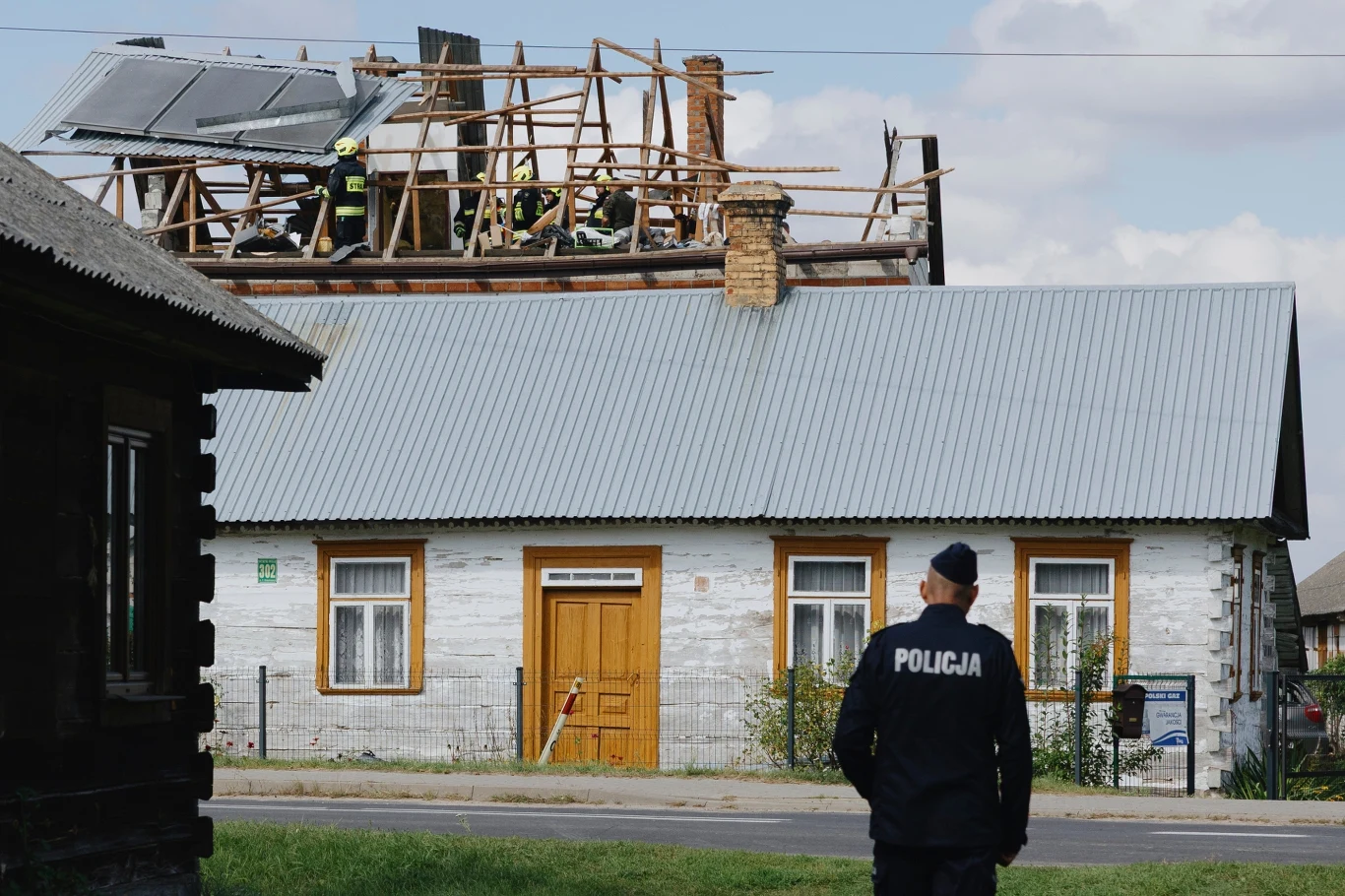 Zniszczony dom w Wyrykach Funkcjonariusz policji obserwuje dom, którego dach jest uszkodzony i naprawiany przez ekipę budowlaną. Na dachu widoczni są strażacy lub pracownicy w uniformach, prowadzący prace remontowe.