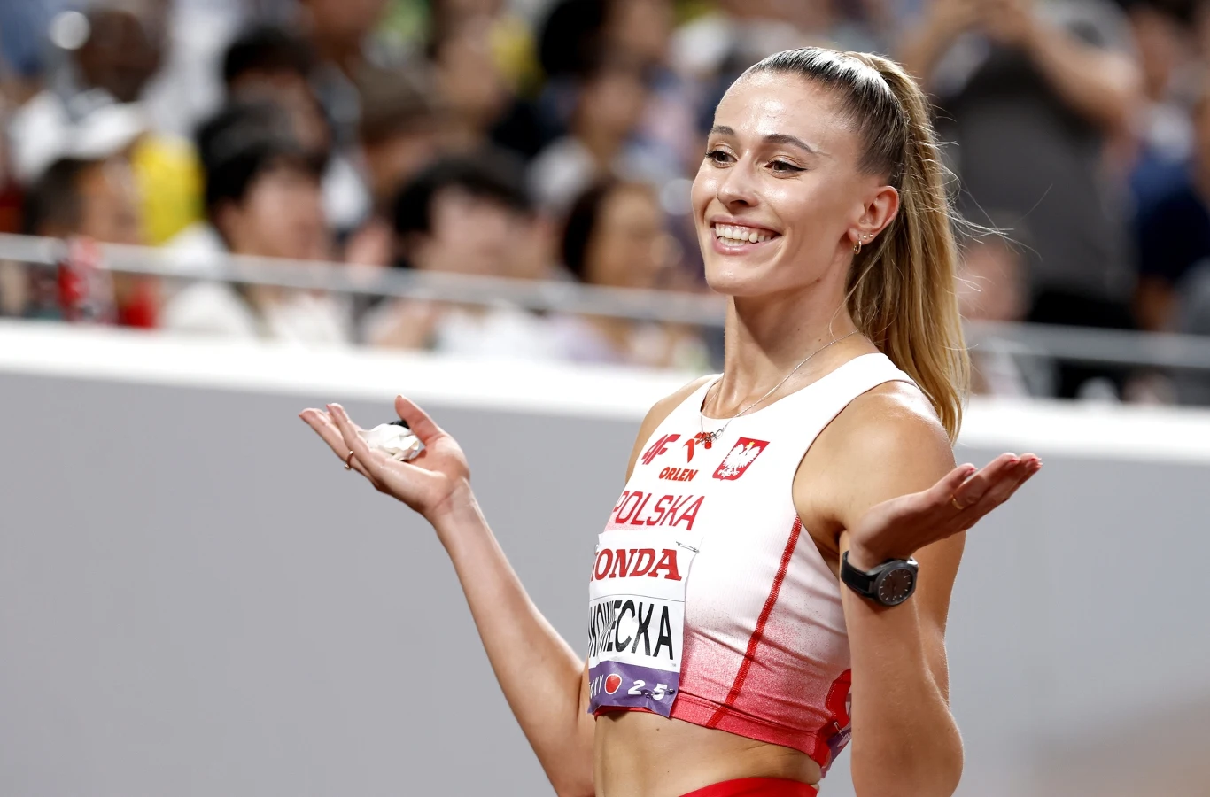 Młoda sportsmenka w stroju reprezentacji Polski lekkoatletycznej uśmiecha się szeroko, unosząc ramiona w geście radości lub zaskoczenia, w tle rozmyta publiczność na stadionie.