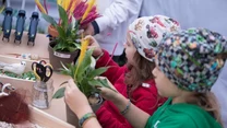 Dwoje dzieci przy stole uczestniczy w warsztatach rękodzielniczych, układając kolorowe rośliny w doniczkach, wokół nich znajdują się narzędzia do prac plastycznych oraz elementy do dekoracji.
