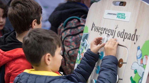 Chłopiec uczestniczy w edukacyjnej zabawie, układając elementy na planszy dotyczącej segregacji odpadów, w tle widoczne inne dzieci.