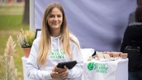 Uśmiechnięta młoda kobieta w białej bluzie z napisem 'Czyste Zielone Miasta' trzyma tablet przy stoisku promocyjnym z ekologicznymi materiałami, obok znajdują się rośliny oraz tekstylia z logo inicjatywy.