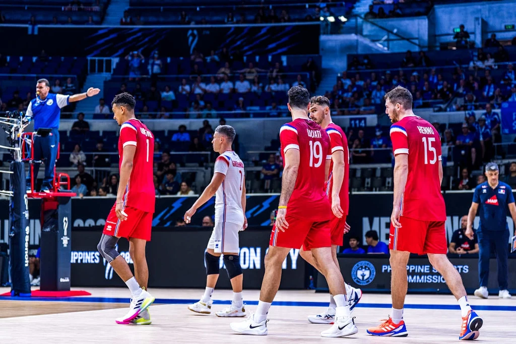 Siatkarze reprezentacji Francji Grupa zawodników siatkarskich w czerwonych strojach znajduje się na boisku w czasie przerwy w meczu, sędzia stoi na podwyższeniu po lewej stronie wskazując kierunek, wokół zawodników widownia oraz elementy hali sportowej.