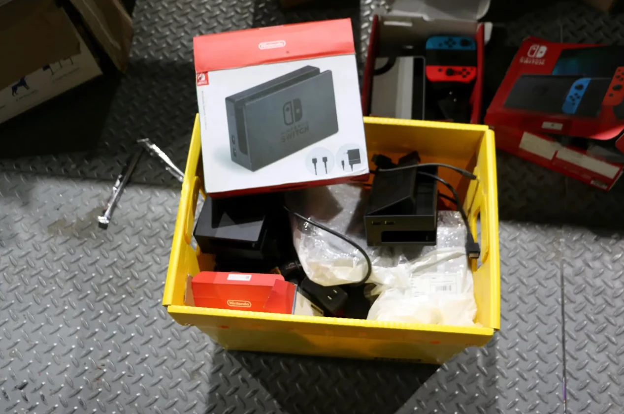 podróbki konsoli Nintendo Żółty pojemnik wypełniony akcesoriami i elementami konsoli Nintendo Switch, wśród nich pudełko z grafiką konsoli oraz zasilacze, przewody i ładowarki. Obok pojemnika kolejne opakowania sprzętu elektronicznego.