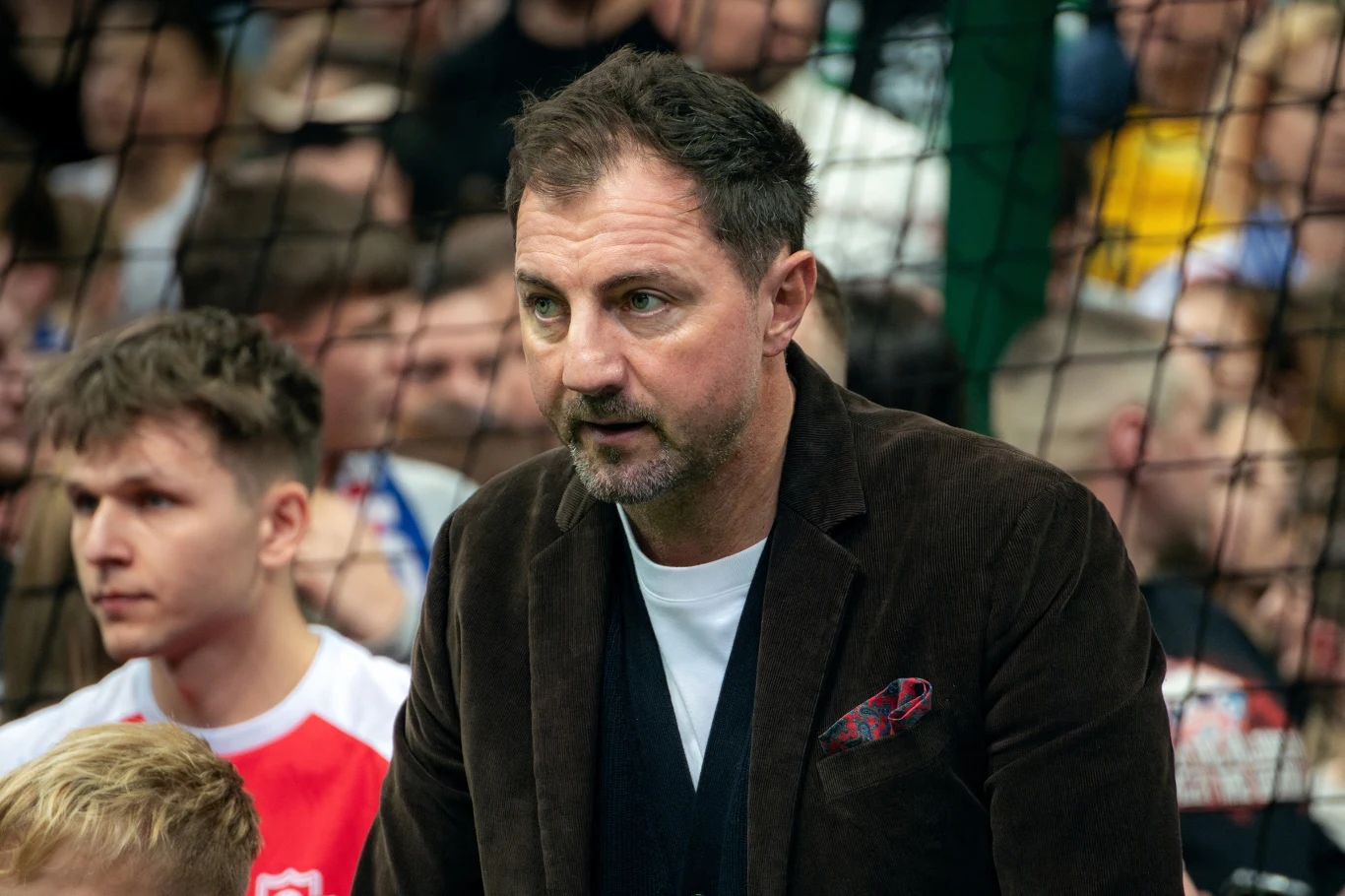 Jerzy Dudek Mężczyzna w ciemnej marynarce stoi na tle siatki, wokół znajdują się inni ludzie w sportowych ubraniach, na twarzy wyraz skupienia.