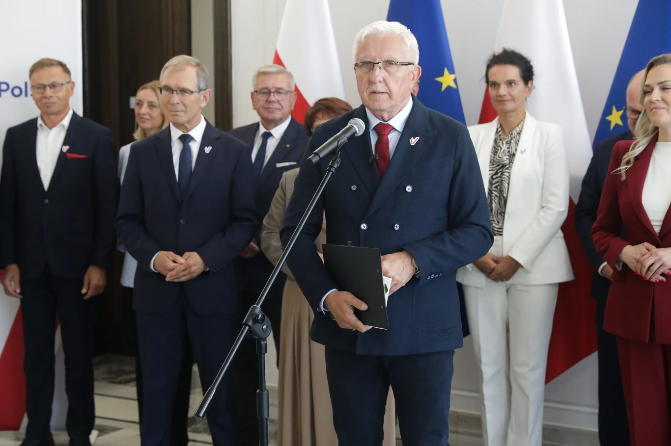 Senatorowie i samorządowcy powołali konserwatywno-liberalną partię - Nowa Polska Grupa dobrze ubranych osób zgromadzona wokół mężczyzny przemawiającego do mikrofonu na tle flag polskiej i unijnej, atmosfera oficjalna, wszyscy ubrani formalnie, wystrój wnętrza nowoczesny