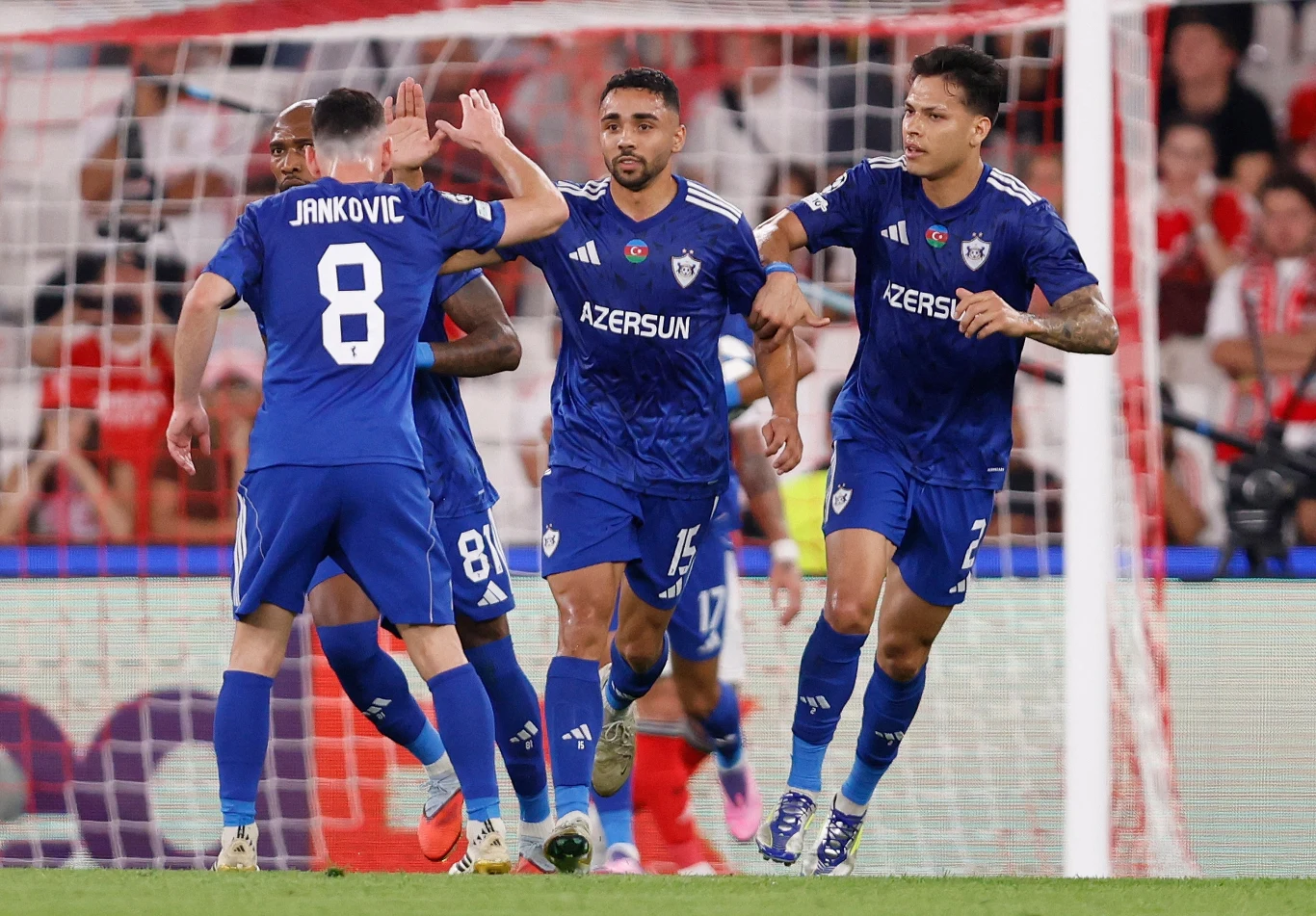 Piłkarze drużyny Qarabağ FK w niebieskich strojach świętują gol, otoczeni przez innych zawodników na boisku podczas meczu piłkarskiego, w tle widoczne trybuny i elementy stadionu.