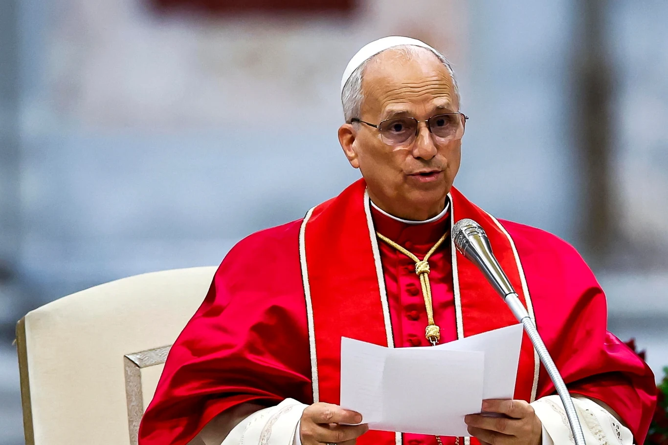Papież Leon XIV Starszy mężczyzna w czerwonym stroju liturgicznym siedzi na fotelu, przemawia do mikrofonu i trzyma kartkę papieru, w tle rozmyte wnętrze kościoła lub katedry.