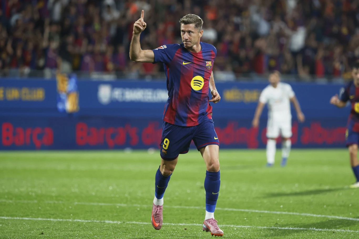 Piłkarz w stroju FC Barcelony z uniesioną ręką świętuje zdobycie gola na boisku, w tle kibice oraz zawodnicy przeciwnej drużyny.