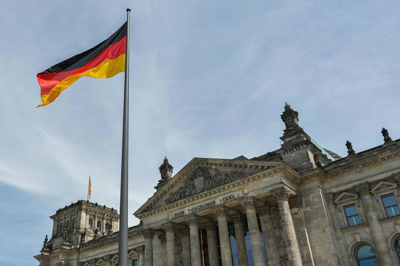 Niemiecka gospodarka pod kreską. Spada produkcja przemysłowa. Zdj. ilustracyjne Budynek niemieckiego Bundestagu z dużą powiewającą flagą Niemiec na pierwszym planie, na tle jasnego nieba.