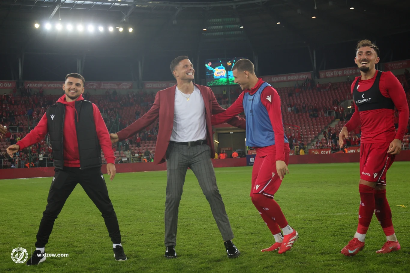 Grupa czterech mężczyzn stojących na murawie stadionu piłkarskiego, uśmiechnięci, ubrani w sportowe stroje i casualowy garnitur, otoczeni światłem reflektorów oraz pustymi trybunami w tle.