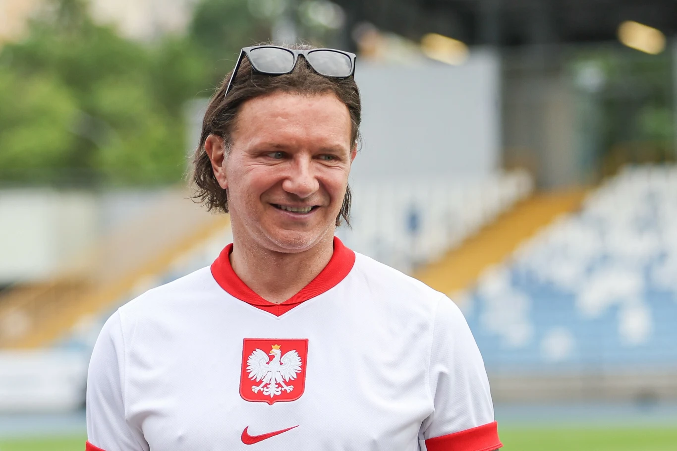 Mężczyzna w koszulce z godłem Polski i logo Nike, uśmiechnięty, z okularami przeciwsłonecznymi na głowie, stoi na tle trybun stadionu sportowego.