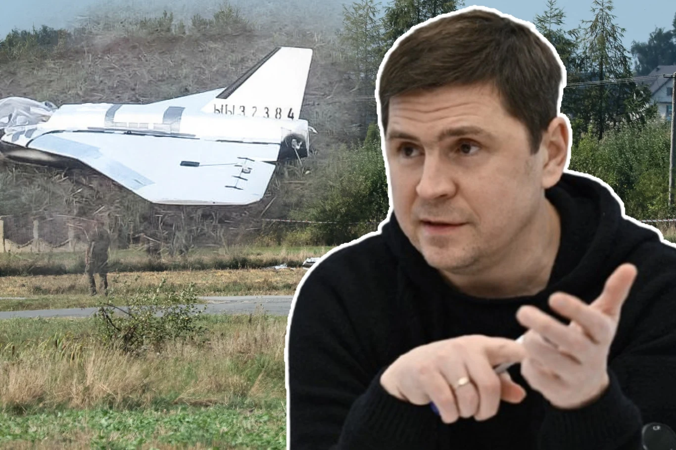 Doradca prezydenta Ukrainy Wołodymyra Zełenskiego Mychajło Podolak Mężczyzna w średnim wieku gestykuluje w kierunku rozmówcy, na tle widoczny wrak samolotu leżący w pobliżu zalesionego terenu, z fragmentem pasma startowego oraz kilku drzew w tle.