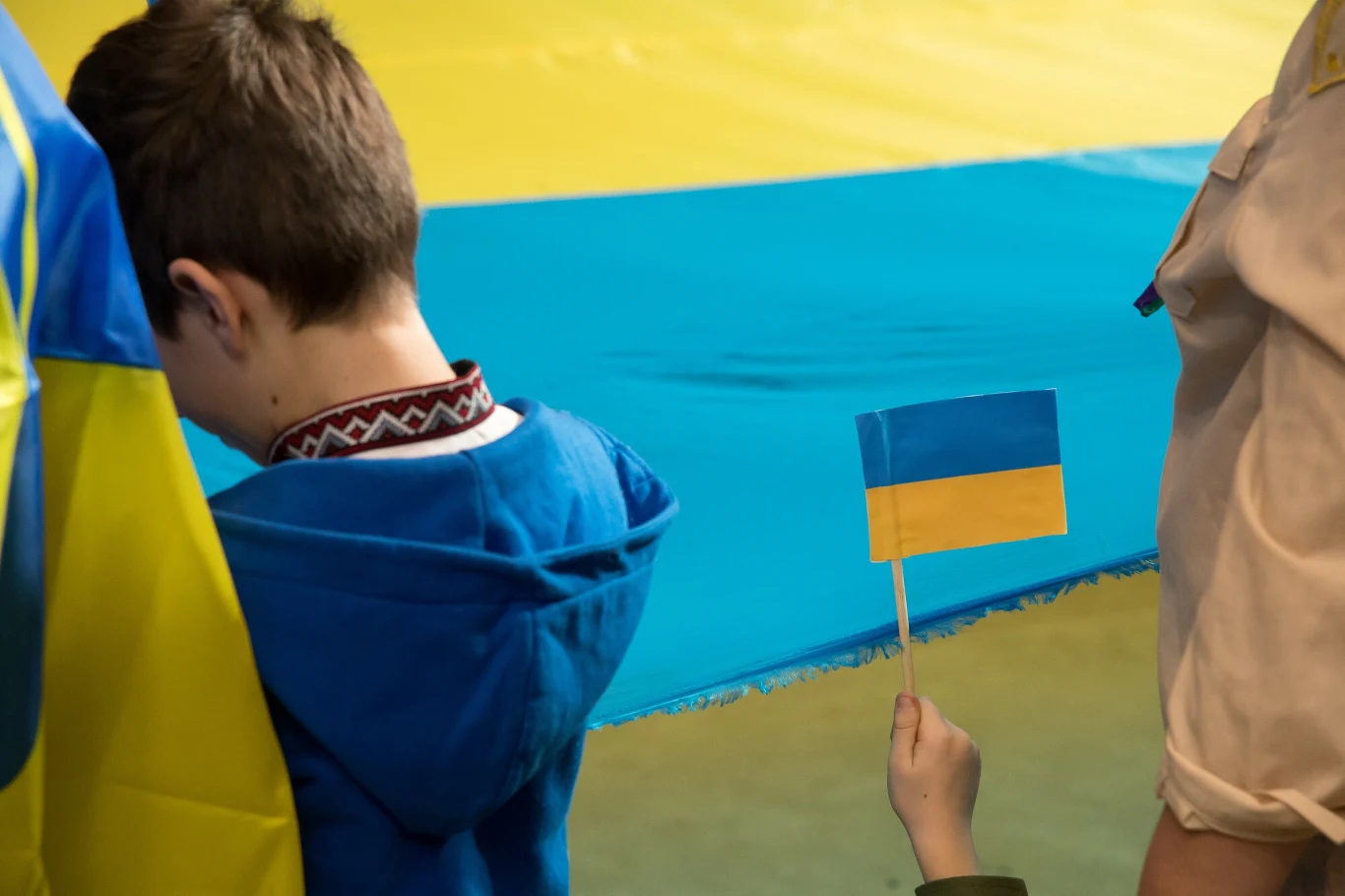 Senat zajął się projektem ustawy o zmianie niektórych ustaw w celu weryfikacji prawa do świadczeń na rzecz rodziny dla cudzoziemców (zdj. ilustracyjne) Chłopiec ze spuszczoną głową stoi w pobliżu dużej flagi Ukrainy, obok uniesiona jest mała ukraińska chorągiewka trzymana przez inną osobę, cała scena utrzymana w kolorach niebieskim i żółtym.