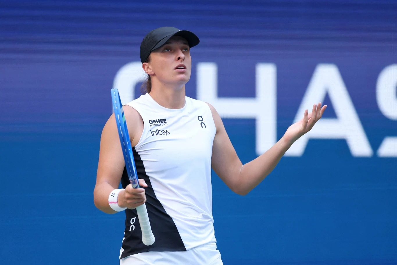 Tenisistka ubrana w sportową białą sukienkę z czarnymi elementami i czapkę z daszkiem trzyma rakietę tenisową na korcie, w geście wyrażającym zdziwienie lub dezaprobatę, w tle widoczna niebieska ściana z niewyraźnymi literami.