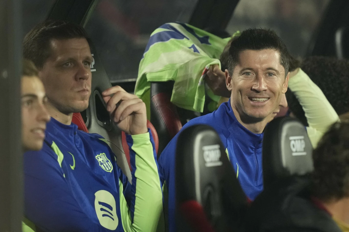 Wojciech Szczęsny i Robert Lewandowski Piłkarze w sportowych strojach siedzący na ławce rezerwowych podczas meczu, jeden z nich uśmiechnięty, drugi skupiony, w tle widoczna bluza przewieszona przez oparcie siedzenia.