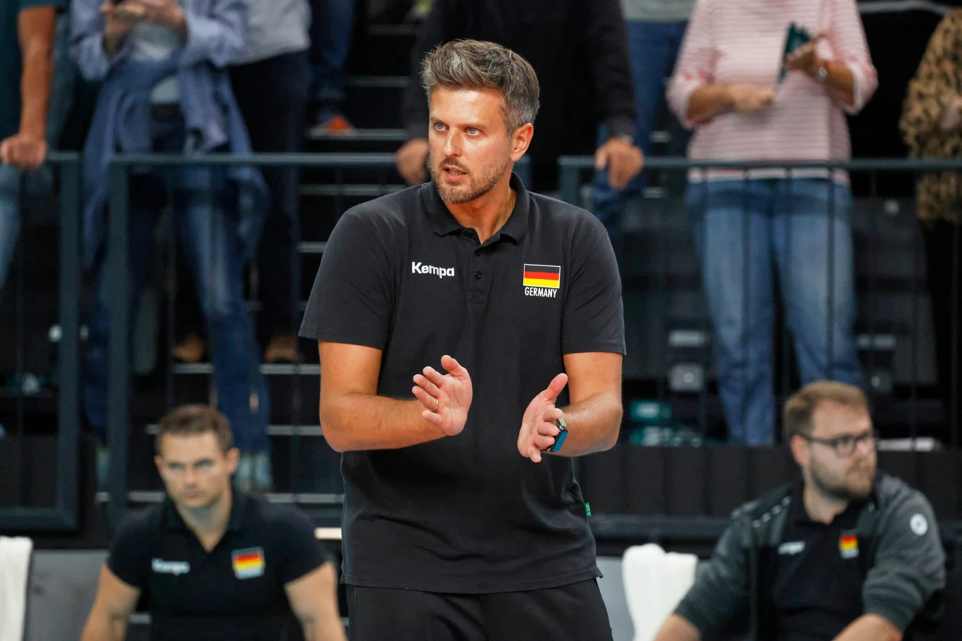 Mężczyzna w czarnej koszulce z flagą Niemiec i napisem Germany stoi na hali sportowej, klaszcząc w dłonie, w tle widzowie oraz inni członkowie zespołu sportowego.