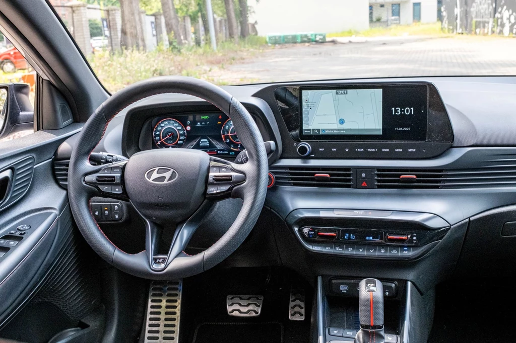 Wnętrze Hyundaia i20 N-Line zdominowane jest przez ekrany. Ale nie brakuje też fizycznych przełączników. Kokpit nowoczesnego samochodu marki Hyundai z cyfrowym panelem wskaźników, dużym ekranem multimedialnym, wielofunkcyjną kierownicą oraz automatyczną skrzynią biegów. W tle widoczne miejsce parkingowe i otoczenie z drzewami.