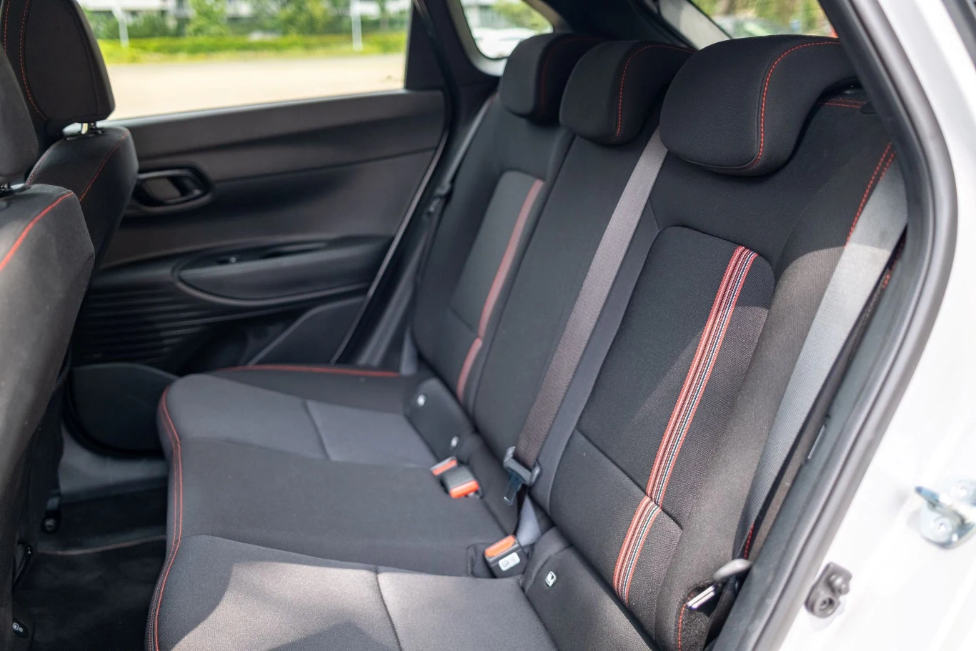 Tylna kanapa samochodu osobowego z trzema miejscami siedzącymi, wykończona tkaniną z kontrastowymi przeszyciami w kolorze czerwonym, pasy bezpieczeństwa na wszystkich miejscach, wnętrze jasne i zadbane.