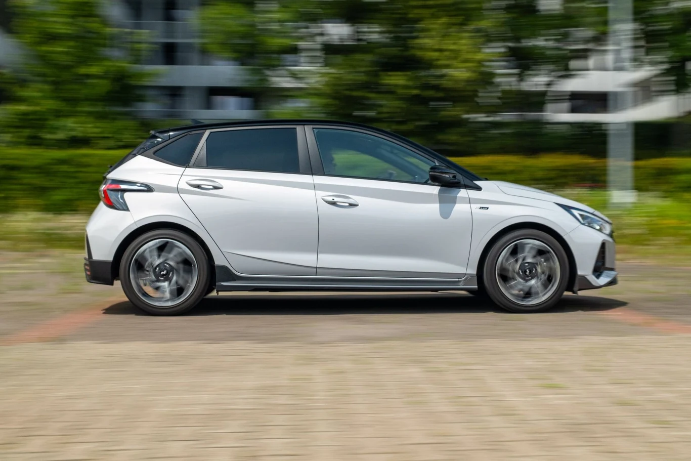 Biały samochód osobowy typu hatchback w ruchu, na tle rozmytego otoczenia miasta z delikatnie widoczną roślinnością, nowoczesny design auta oraz aluminiowe felgi dominantują na pierwszym planie.