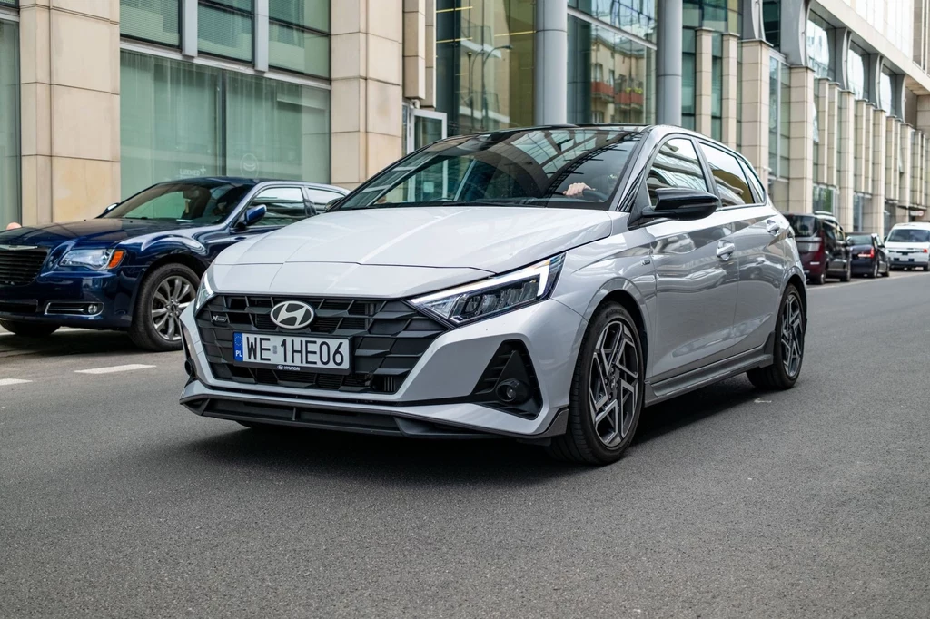 Średnie zużycie paliwa Hyundaia i20 N-Line w trasie może wynieść nawet 7,6 l/100 km. Srebrny samochód Hyundai stoi na ulicy przed nowoczesnym budynkiem, w tle widoczne są inne zaparkowane pojazdy i przeszklona fasada biurowca.
