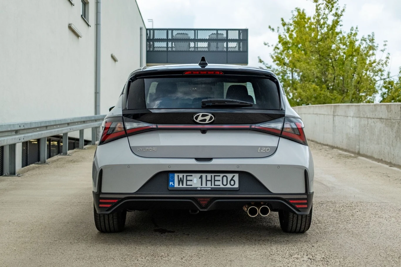 Tylna część jasnoszarego samochodu marki Hyundai i20 zaparkowanego na betonowym podjeździe, z widoczną polską tablicą rejestracyjną oraz charakterystycznym designem świateł i podwójnym wydechem.