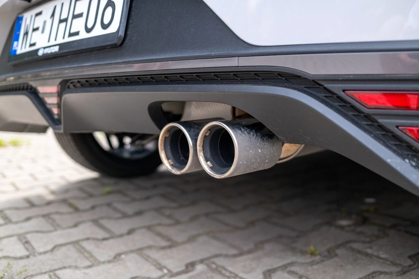 Podwójna rura wydechowa samochodu osobowego zaparkowanego na kostce brukowej, tylny zderzak w kolorze czarnym i fragment tablicy rejestracyjnej.