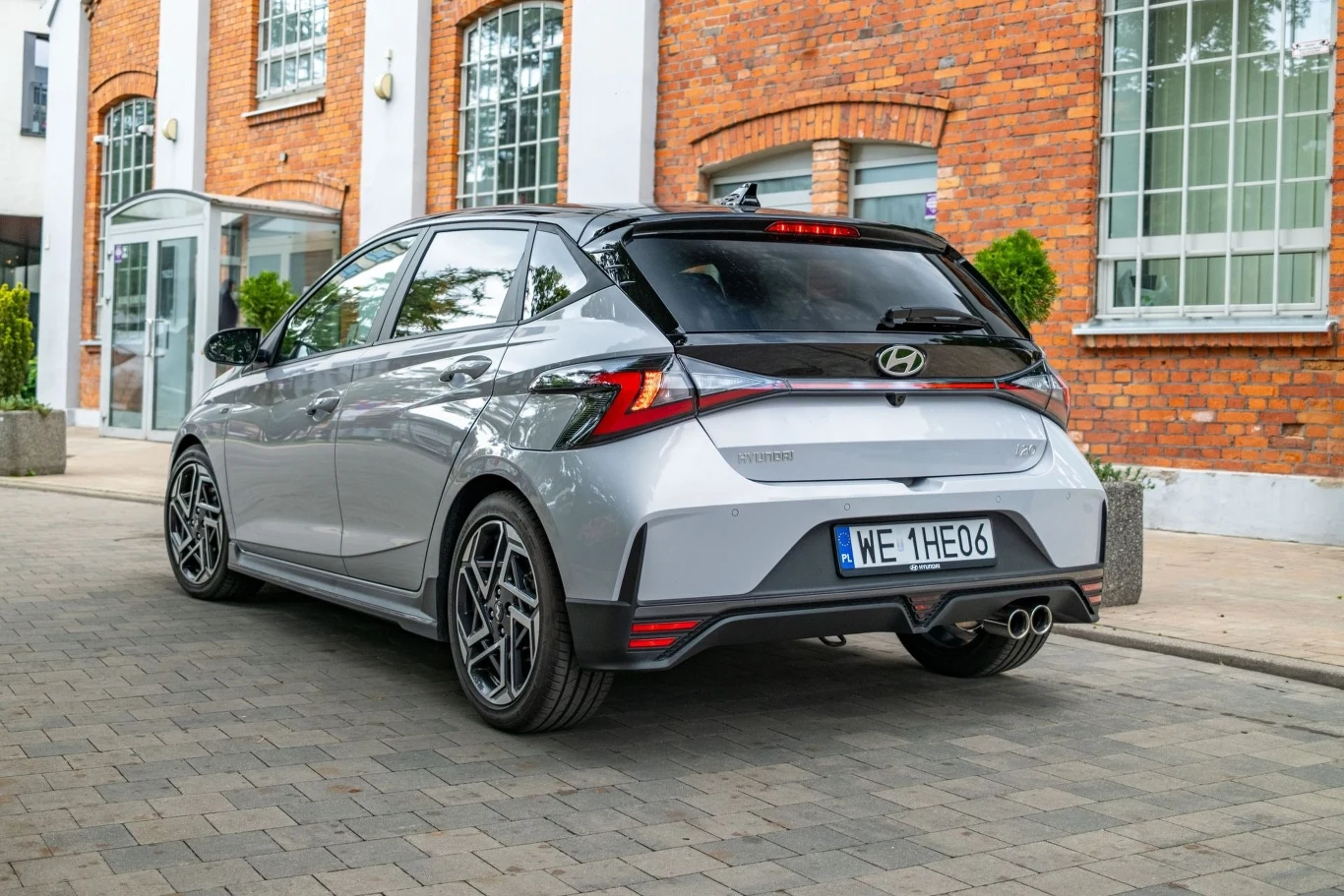 Srebrny samochód osobowy Hyundai i20 N stoi zaparkowany na brukowanej ulicy przy nowoczesnym budynku z ceglanymi ścianami i dużymi oknami.