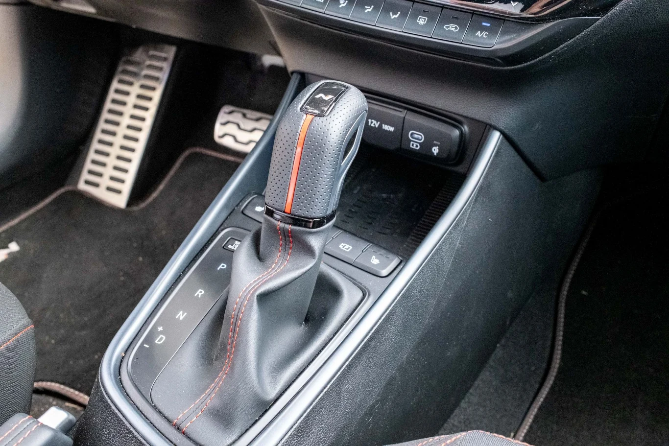 Dźwignia automatycznej skrzyni biegów z czarną skórzaną wykładziną i pomarańczowym przeszyciem obok panelu centralnego samochodu, widoczne również pedały gazu i hamulca.