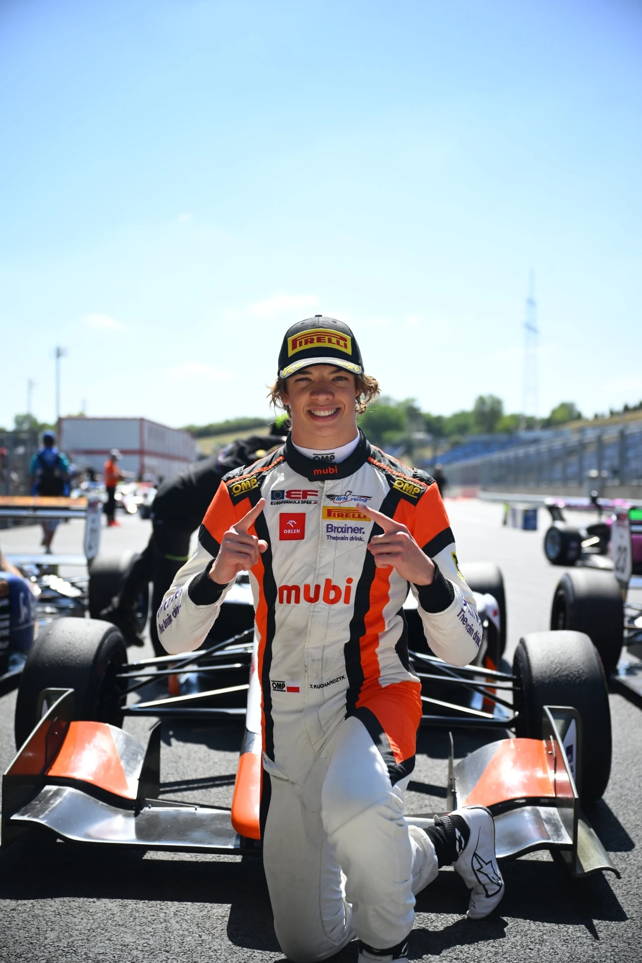 Młody kierowca wyścigowy w kombinezonie z logotypami sponsorów klęczy przed bolidem na torze, trzyma dwie karty i uśmiecha się szeroko, w tle widoczna jest linia startowa oraz inni zawodnicy przy samochodach.