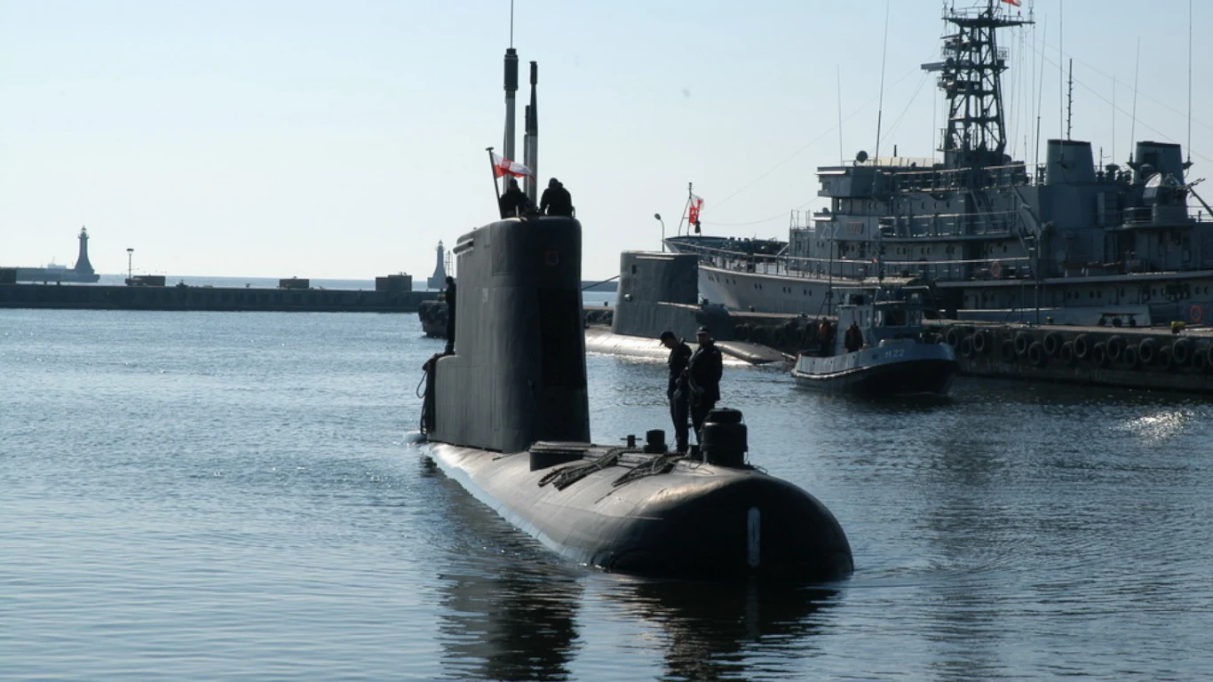 Okręt podwodny wynurzający się w porcie, na jego pokładzie kilku członków załogi, w tle widoczne inne jednostki wojskowe i infrastruktura portowa.