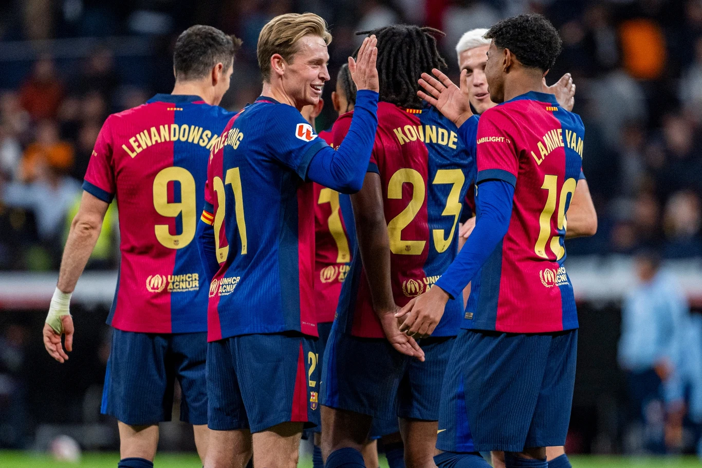 Zawodnicy FC Barcelona Grupa piłkarzy w czerwono-niebieskich strojach świętuje wspólnie zdobycie gola na boisku, kilku zawodników przybija sobie piątki, na koszulkach widoczne nazwiska i numery sportowców.