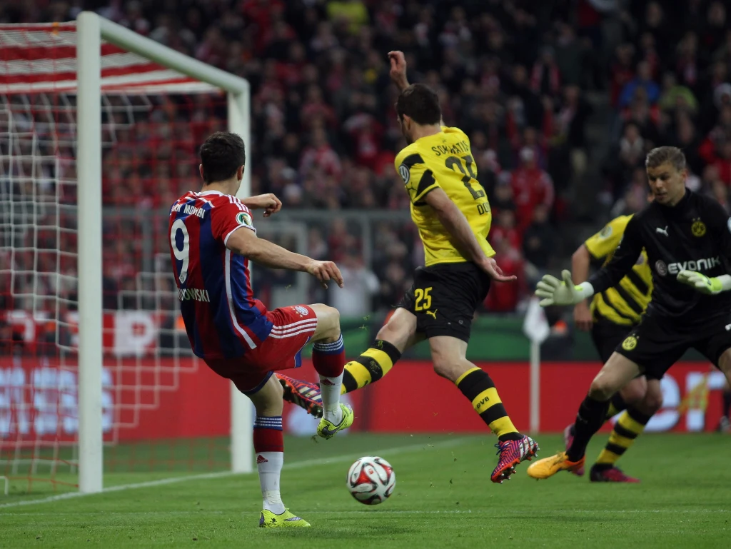 Kadr z meczy Bayern - Borussia (28 kwietnia 2015) Piłkarz w czerwono-białym stroju próbuje zdobyć bramkę, oddając strzał z bliskiej odległości, naprzeciwko niego znajduje się obrońca w żółto-czarnym stroju, a bramkarz przygotowuje się do obrony, na tle widoczne trybuny pełne kibiców.