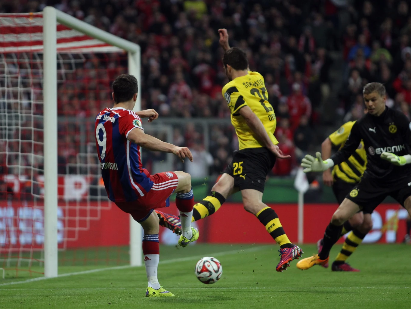 Piłkarz w czerwono-białym stroju próbuje zdobyć bramkę, oddając strzał z bliskiej odległości, naprzeciwko niego znajduje się obrońca w żółto-czarnym stroju, a bramkarz przygotowuje się do obrony, na tle widoczne trybuny pełne kibiców.