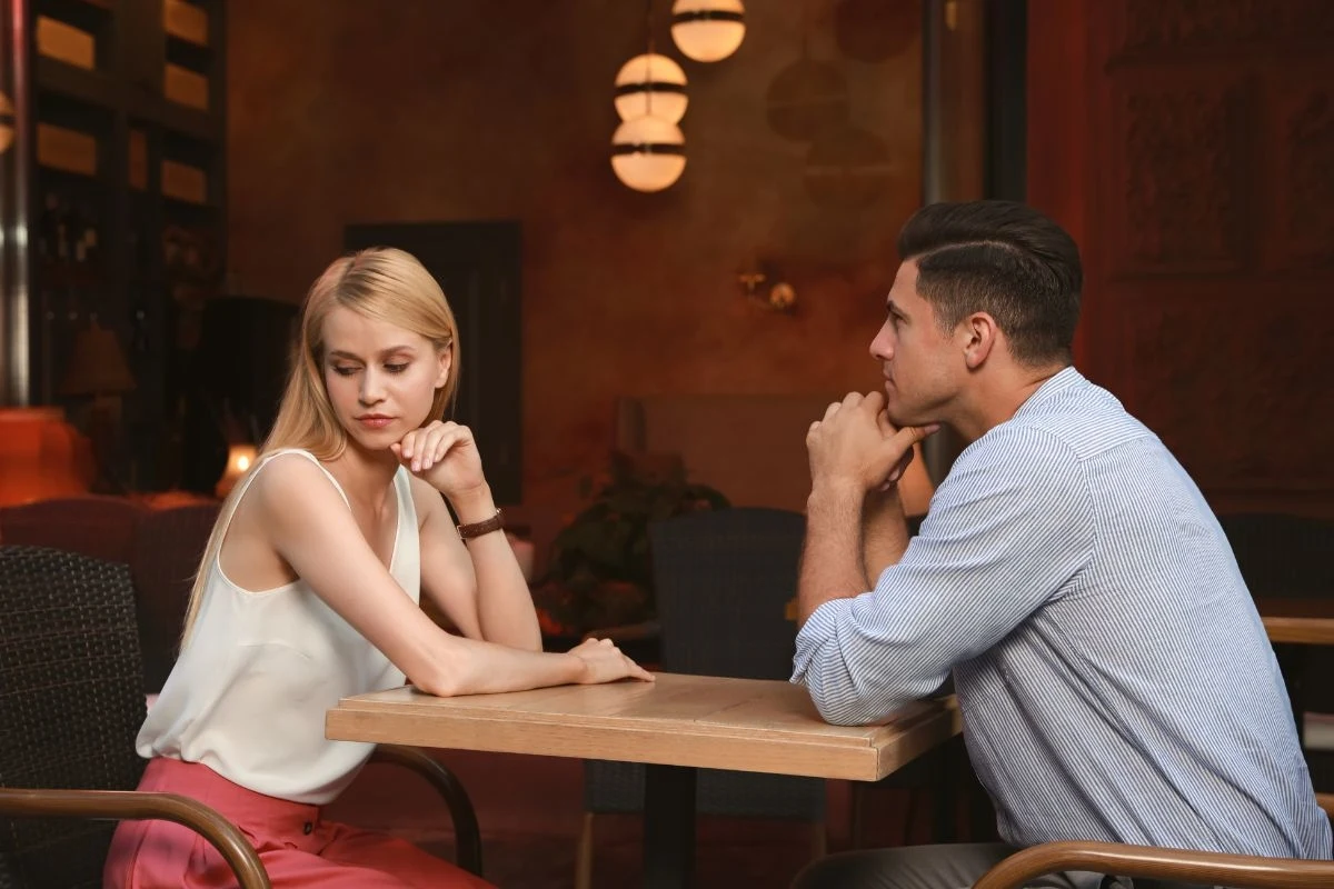 Para siedzi naprzeciw siebie przy stoliku w przyciemnionej restauracji, kobieta odwraca wzrok i wydaje się być zamyślona lub zmartwiona, mężczyzna patrzy na nią z poważnym wyrazem twarzy, oboje wspierają głowy na rękach, atmosfera jest napięta i pełna ...