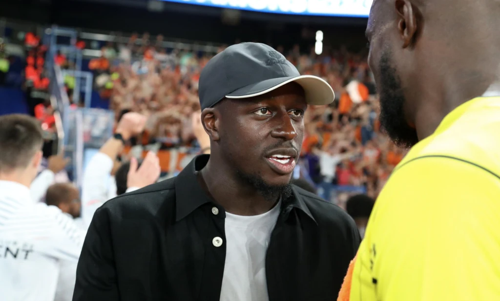 Mężczyzna w czapce z daszkiem i czarnej kurtce prowadzi rozmowę z innym mężczyzną w żółtej koszulce, w tle tłum kibiców na stadionie sportowym.