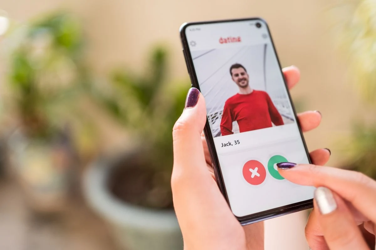 Ekran telefonu z otwartą aplikacją randkową, na którym widoczny jest profil mężczyzny w czerwonym swetrze o imieniu Jack i wieku 35 lat, palec wskazujący na przycisk wyboru.