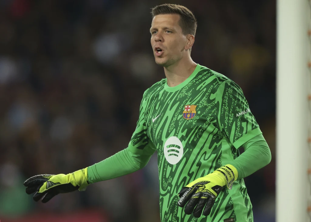 Wojciech Szczęsny Piłkarz ubrany w zielony strój bramkarski z logo klubu piłkarskiego i rękawicami, stojący na tle boiska podczas meczu, z widocznym gestem komunikacyjnym.