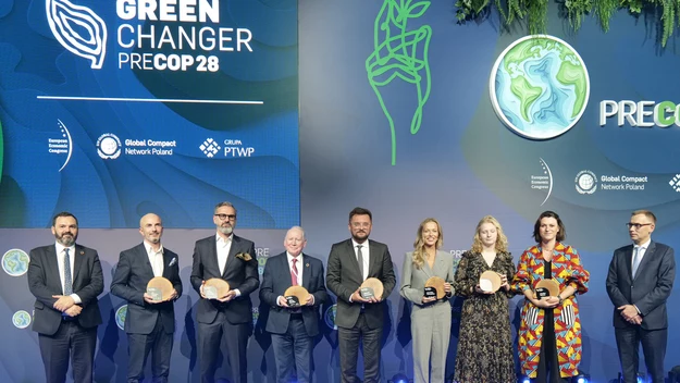 Grupa ośmiu osób ubranych oficjalnie stoi na tle ściany z logo wydarzenia Green Changer PRECOP28, trzymając w dłoniach nagrody, otoczona przez profesjonalne oświetlenie sceniczne.