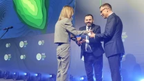 Trzy osoby w formalnych strojach na scenie podczas oficjalnego wydarzenia, jedna osoba wręcza nagrodę lub wyróżnienie drugiej, w tle grafika i logo konferencji PreCOP28, całość oświetlona lampami scenicznymi.